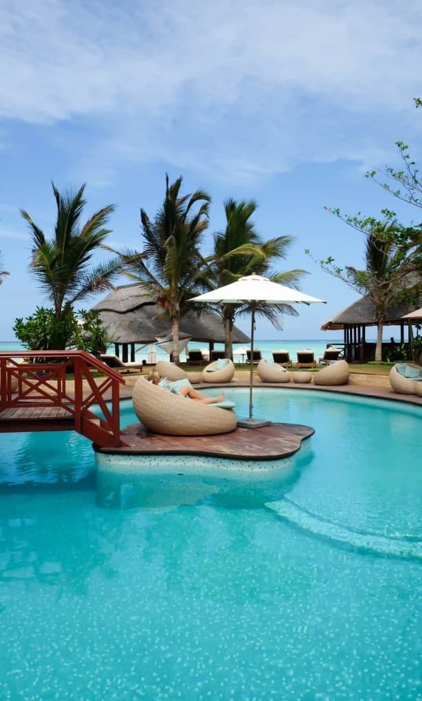 Tropical resort lagoon pool with rattan loungers, parasols and ocean views through swaying palms in Zanzibar.