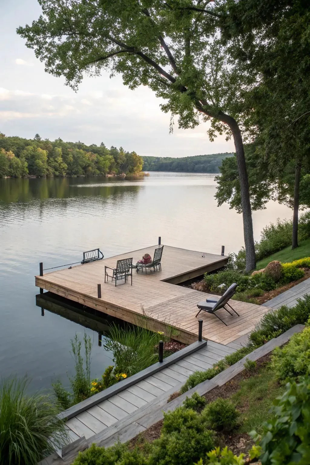 Timber lake deck with lounge seating reaching over glassy water surrounded by forested shoreline at dusk.