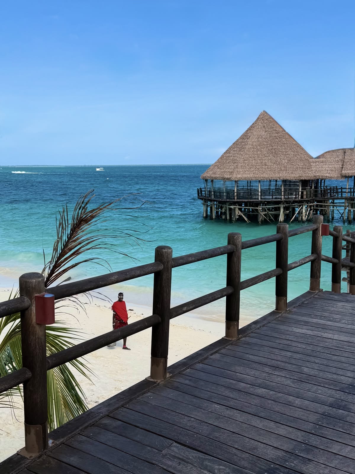 Wooden jetty leading to a thatched overwater restaurant above turquoise water as a Maasai walks the white-sand beach in Zanzibar.